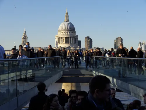 The image is an urban photograph taken in London, United Kingdom, during a sunny late afternoon in early spring. The main subject is the Millennium Bridge, a pedestrian bridge that spans the River Thames, with many people walking across it. Prominently featured in the background is the dome of St Paul's Cathedral, a well-known church and architectural landmark in London. The cathedral’s impressive architecture dominates the skyline, further emphasizing the city setting. Surrounding St Paul's Cathedral are several other notable buildings, contributing to the overall urban landscape. The composition highlights the relationship between modern bridge architecture and the historic cathedral, capturing a vibrant moment within the heart of the city.